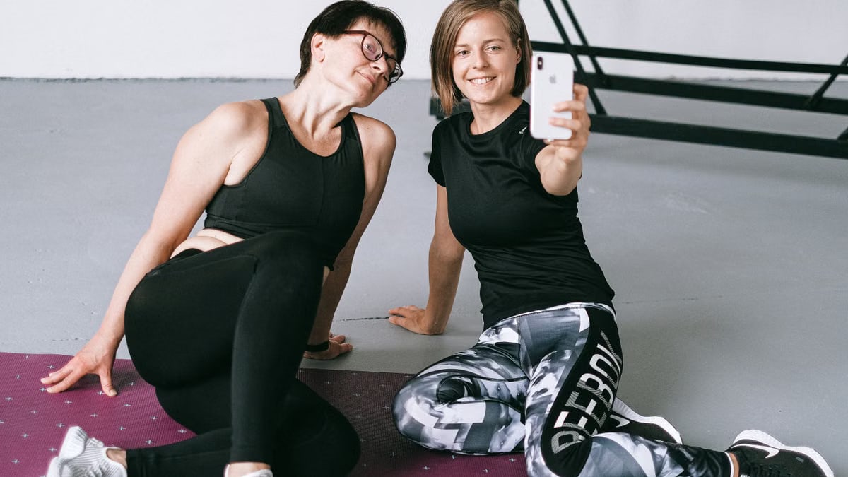 Two women in athletic clothing sitting on a yoga mat indoors, capturing a selfie post workout.
